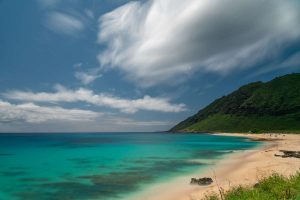 An uncrowded beach in Hawaii with green water, blue sky, and white clouds